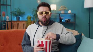 Young man sits on couch eating popcorn and watching interesting TV serial sport game online at home video