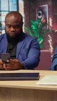 Young adults in their living space browsing webpages on smartphones together while they sit at the desk, having fun and enjoying leisure time with their gadgets. Cozy apartment. Camera A. video