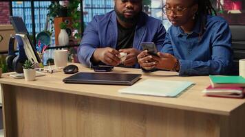 Young black couple sharing ear buds to listen to webinar on smartphone, working together on their freelancing tasks at home. Man and woman discussing about a work project. Camera A. video