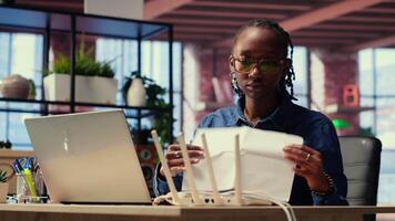 Black woman at home examines the modem and troubleshooting guide book, struggling to fix Wi Fi router and restore her internet connection for remote freelance tasks. Signal loss. Camera B. video