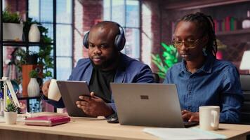 Black woman is working while her partner listens to music next to her, enjoying their time at home together. Freelancer solving her daily tasks on laptop, boyfriend scrolling web. Camera B. video