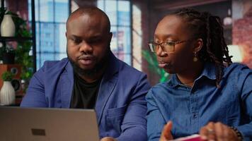 Black remote employees organizing calendar appointments using laptop and ledger, checking papers and synchronizing important tasks on schedule. Man and woman review files at home. Camera A. video
