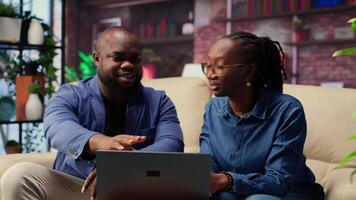Black couple in living room discusses creative ideas for an online business, using modern technology to research trends and improve efficiency. Motivation and support for success. Camera B. video