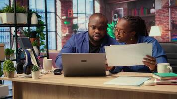 Young couple evaluating monthly reports numbers with a laptop and papers, analyzing figures and calculating the balance before deadline. African american people working from home. Camera A. video