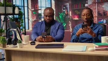 Young adults in their living space browsing webpages on smartphones together while they sit at the desk, having fun and enjoying leisure time with their gadgets. Cozy apartment. Camera A. video