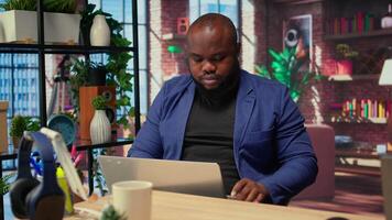 Teleworker at home office editing documents on his personal laptop, submitting reports and files before deadline. Black freelancer managing digital paperwork within the work platform. Camera A. video