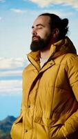 Vertical Hiker dressed in warm clothing stands on a mountain peak after climbing to the top, gazing at the breathtaking landscape of rugged hills and vast sky. Adventure with scenic view. Camera B. video