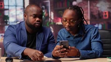 Young black couple sharing ear buds to listen to webinar on smartphone, working together on their freelancing tasks at home. Man and woman discussing about a work project. Camera B. video