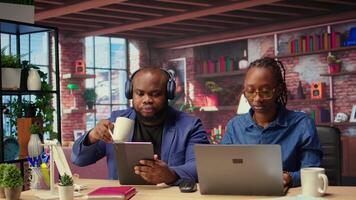 Black woman is working while her partner listens to music next to her, enjoying their time at home together. Freelancer solving her daily tasks on laptop, boyfriend scrolling web. Camera A. video