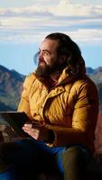 Vertical Camper fighting the strong wind while he uses a tablet at a high altitude campsite, searching for better signal and risking his safety. Dangerous mountain range cold weather. Camera B. video