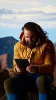 Vertical Male hiker struggles to work online in windy conditions on a remote mountain peak. Battling weak signals and cold weather, explorer is obsessed with technology and the internet. Camera B. video