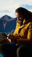 Vertical Camper fighting the strong wind while he uses a tablet at a high altitude campsite, searching for better signal and risking his safety. Dangerous mountain range cold weather. Camera A. video