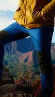 Vertical Hiker tourist with backpack on top of mountain puts foot on the rocks stone and enjoys climbing success. Young man looks at picturesque valley scenic landscape, freedom. Camera A. video