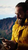 Vertical Nature lover relaxing on mountain top getting online with his phone at a scenic viewpoint, looking out over the landscape. Hiker staying always online and connected, panoramic scenery. Camera A. video