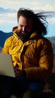 Vertical Mountain adventurer prioritizes his online business over safety, working on his laptop despite the extreme winds. The breathtaking landscape is overshadowed by his internet dependency. Camera A. video