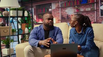Black man and woman sit together in their cozy apartment space, discussing digital marketing strategy on a laptop. Supporting each other in their remote work and freelancing journey. Camera A. video
