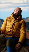 Vertical Male mountain hiker celebrates a victorious climb and resting at the top, arms lifted in excitement while he sits on the rocks. Panoramic view and crisp air in a scenic background. Camera B. video
