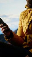 Vertical Traveler sitting and browsing his phone on the rocky mountain top, feeling pleased with the stunning mountain range and scenic landscape. Obsessed with technology and internet. Camera B. video
