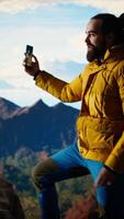 Vertical Male content creator recording a surrounded by mountain peaks in the clouds, climbing a mountain range hill and live streaming for his social media audience. Adventurous spirit. Camera A. video