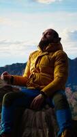Vertical Male mountain hiker celebrates a victorious climb and resting at the top, arms lifted in excitement while he sits on the rocks. Panoramic view and crisp air in a scenic background. Camera A. video