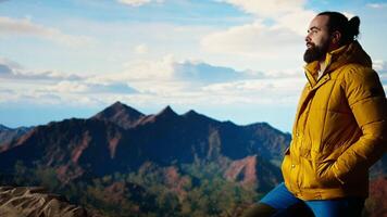 Hiker dressed in warm clothing stands on a mountain peak after climbing to the top, gazing at the breathtaking landscape of rugged hills and vast sky. Adventure with scenic view. Camera A. video