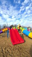 criança slides às Parque infantil. uma criança slides baixa uma vermelho deslizar às uma ensolarado Parque infantil preenchidas com colorida jogar equipamento e azul céus. video