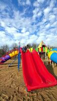 Kid on bright slide. A young child joyfully slides down a vibrant red slide at a playground surrounded by bright structures and blue skies. video