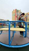 Kind liebt Park Spielplatz Fahrt. ein jung Junge im ein bunt Jacke steht auf ein Spinnen Spielplatz Fahrt, lächelnd während umgeben durch modern Gebäude. video