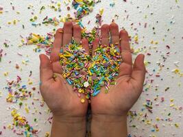 Child's hands are covered with multi-colored sugar sprinkles and form a heart shape. Child plays with small sprinkles topping. photo