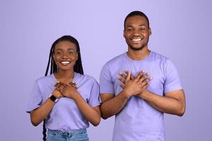 Thank You. Grateful Young Black Couple Holding And Pressing Hands To Chest Expressing Gratitude Posing Over Purple Violet Studio Background And Looking At Camera. Appreciation Concept photo