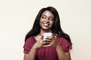 Pretty long-haired young black woman with newest cellphone looking at copy space over beige studio background, using nice dating, entertaining mobile application on smartphone, panorama photo