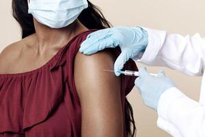 Medical worker wearing gloves and protective face mask making vaccination against coronavirus in shoulder for young black lady, beige studio background, closeup. Safety vaccination concept photo