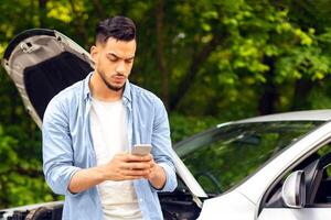 Sad arab man looking for nearest auto service, using smartphone, got broken at highway on a way to countryside. Middle-eastern guy chatting with friends, asking for help with auto photo