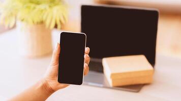 Technology Concept. Unrecognizable afro lady holding and showing smartphone with black blank screen for mock up. Laptop computer with present box or delivery package on keyboard in blurred background photo