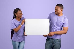 Cheerful Black Couple Holding And Showing Blank Paper Poster For Your Text Smiling Looking At Placard Standing Isolated On Purple Violet Studio Wall. Spouses With White Square Board For Advertising photo