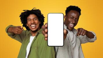 Two Black Male Friends Demonstrating Smartphone With Blank White Screen And Pointing On It, Happy Young African American Guys Recommending New Mobile App Or Website, Cropped Image, Mockup photo