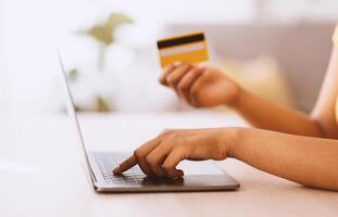 Online Shopping Concept. Side view of unrecognizable african american woman holding credit card in hand and using laptop, ordering gifts, typing on keyboard, blurred background, selective focus photo