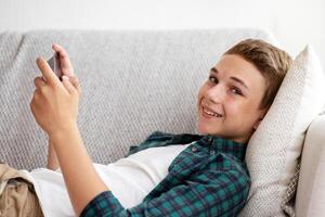 Side view of positive teen guy gaming on smartphone while resting at home photo