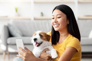 Attractive young asian woman holding fluffy puppy and taking selfie on smartphone while enjoying time together at home, happy jack russel terrier owner taking photos with dog, copy space
