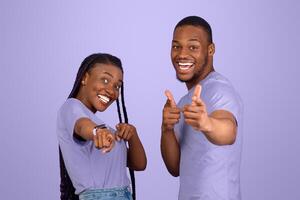 Hey, You. Portrait of cheerful African American guy and lady pointing two index fingers at camera, standing isolated on purple violet studio background. Positive young friends choosing and indicating photo