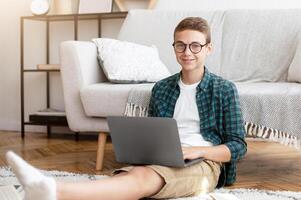 Cheerful male teenager with glasses doing homework, sitting on floor at his room, using books and laptop, empty space photo