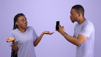 Another Life Online, Cheating Concept. Angry black man checking his girlfriend mobile phone, showing provocative messages and asking for explanation, lady spreading hands, violet studio background photo