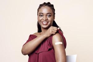 Happy black lady sitting on chair, showing adhesive medical band on her shoulder after vaccination and smiling at camera, beige studio background, copy space. Immunization during COVID-19 pandemic photo