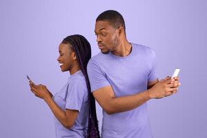 Profile side view portrait of curious black guy trying to look at his girlfriend's smartphone, smiling lady holding using gadget and chatting, couple standing back to back, violet purple background photo