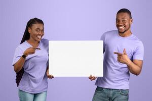 Portrait of cheerful smiling black couple holding and pointing fingers at white paper placard for advertisement or text, free copy space for advertising, isolated on violet purple studio background photo