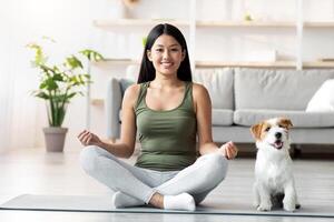 Athletic asian young woman doing yoga with her adorable loyal red and white doggy at home, chinese lady sitting in lotus pose, jack russel puppy sitting by owner on fitness mat, copy space photo