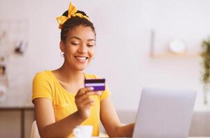 Online Wallet And Banking Concept. Smiling black woman using laptop and credit card, making financial transaction, sitting at desk, ordering purchases, checking debit card number and security code photo