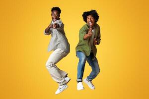 Two Cheerful Black Guys Pointing At Camera While Jumping Over Beige Studio Background, Emotional Young African American Males Saying Gotcha, Having Fun Together, Full Length Shot, Copy Space photo