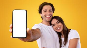 Using Cool Application. Portrait of cute millennial couple holding smartphone with white screen in hand, recommending mobile app, advertising your website, hugging, mock up for design. Selective focus photo