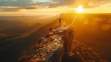 A person stands triumphantly on the edge of a rocky cliff, arms stretched wide, as the sun sets over a vast mountainous landscape, casting warm golden hues. photo